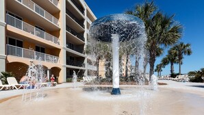 A water fountain with multiple jets, surrounded by a pool, palm trees, and a multi-story building with balconies. - Ocean Front 3 BR/3BA/Ground Floor walk out Updated (Fort Walton Beach)