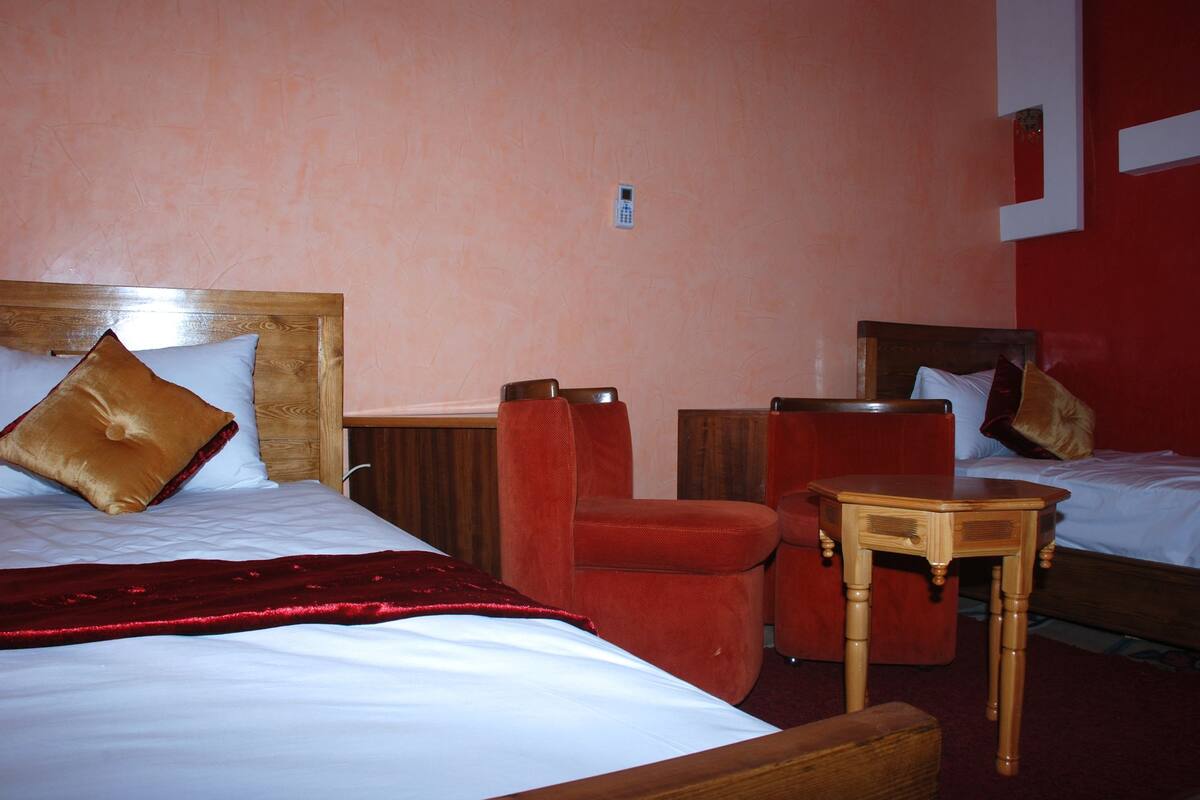 A hotel room with two beds, a wooden headboard, a red armchair, a small wooden table, and a telephone on the wall.