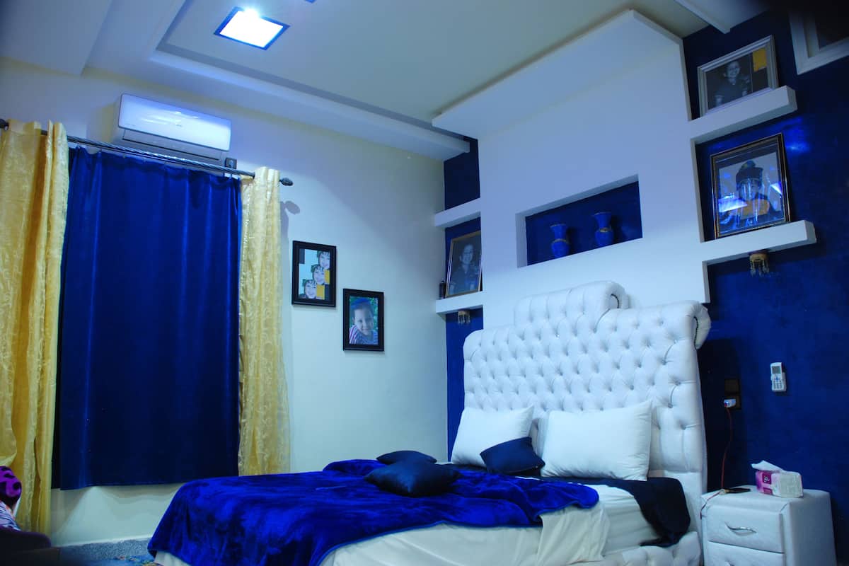 A bedroom with a large bed, a bedside table, a wall-mounted air conditioner, and framed pictures on the wall.