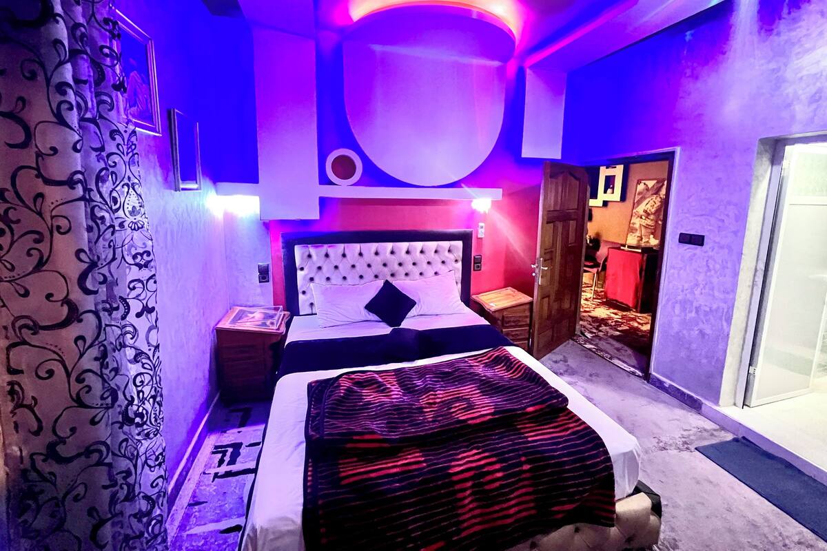 A modern bedroom with a tufted headboard, a bed with a patterned blanket, a bedside table with a framed picture, and a door leading to another room.