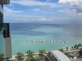 Vista de varanda sobre um mar tranquilo, com vários barcos, céu claro e uma linha de costa distante com palmeiras.