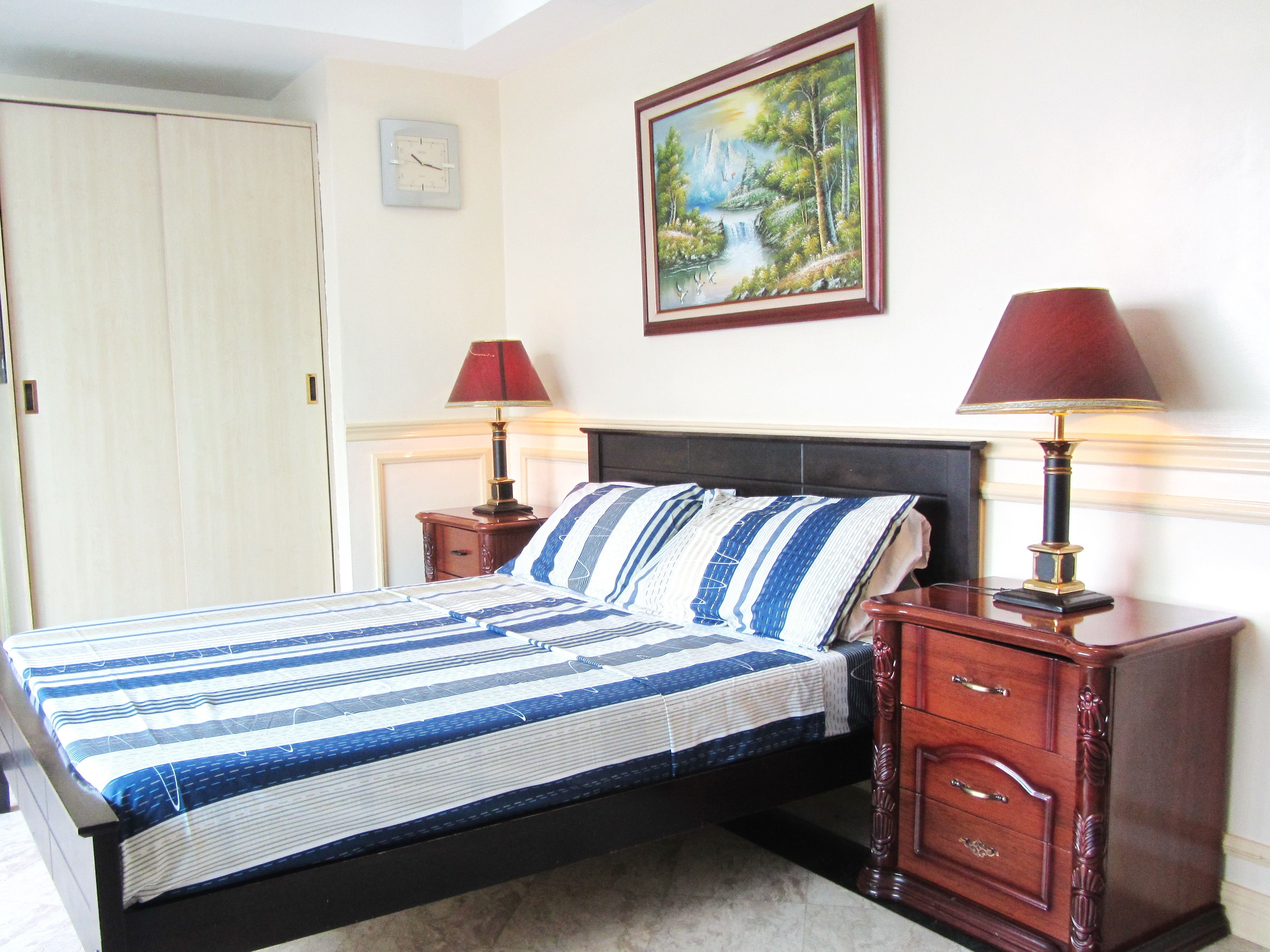 A neatly made bed with striped bedding, two bedside tables with lamps, a clock on the wall, and a framed painting above the bed.