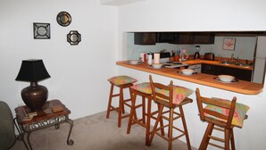 Una cocina compacta con barra, sillas de madera y una mesita auxiliar con lámpara.