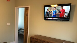 A bedroom with a TV displaying a sports event, a wooden dresser, a bed, and a door leading to another room. - 1st Class Rentals Munson Manor IV  at Columbia Street (Oneonta)