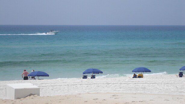 Une plage de sable blanc, avec des parasols bleus et un bateau sur l’eau.
