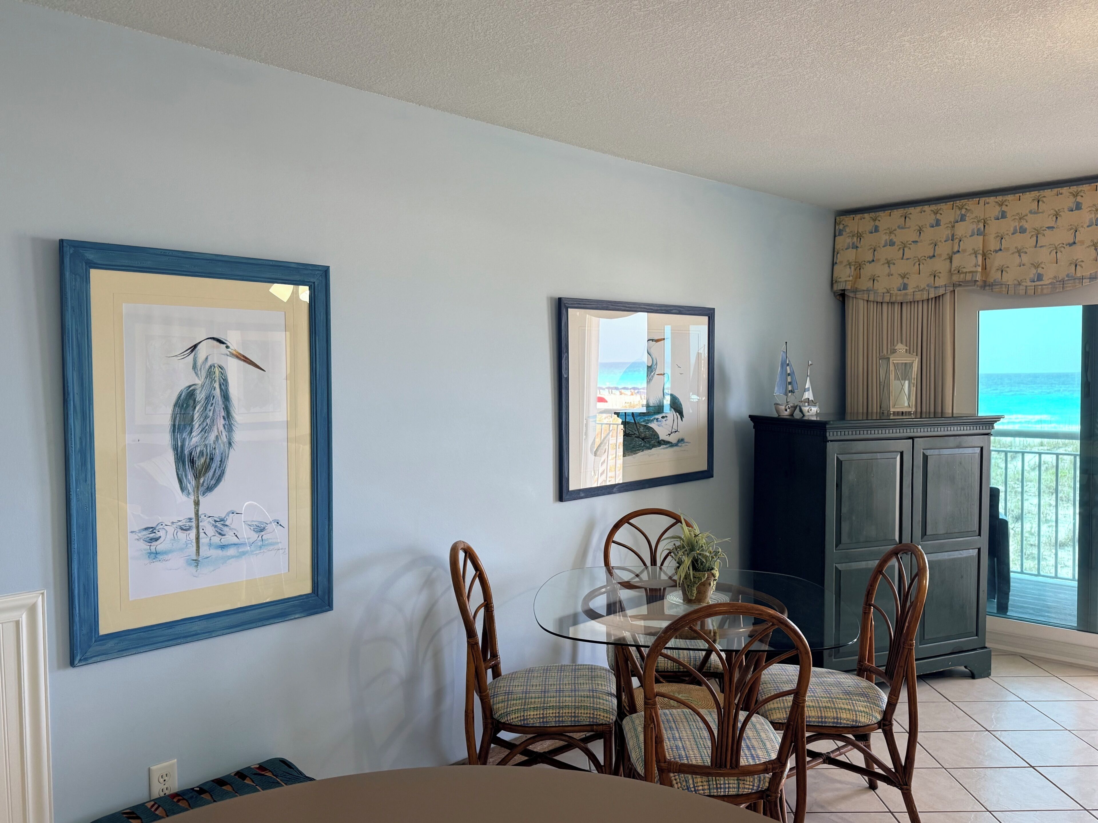 A dining area with a glass table and wicker chairs, framed artwork on the walls, and a view of the ocean through a balcony.