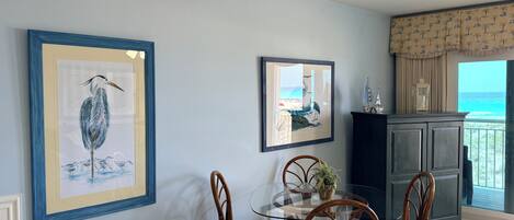 A dining area with a glass table and wicker chairs, framed artwork on the walls, and a view of the ocean through a balcony.