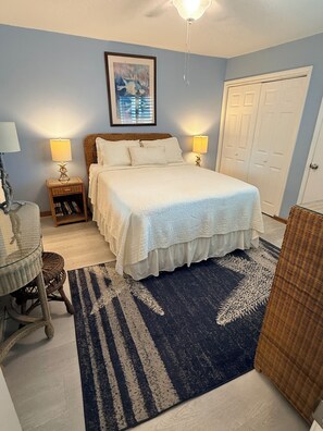 A bedroom with a bed, two bedside lamps, a framed picture on the wall, a door, and a wicker chair.