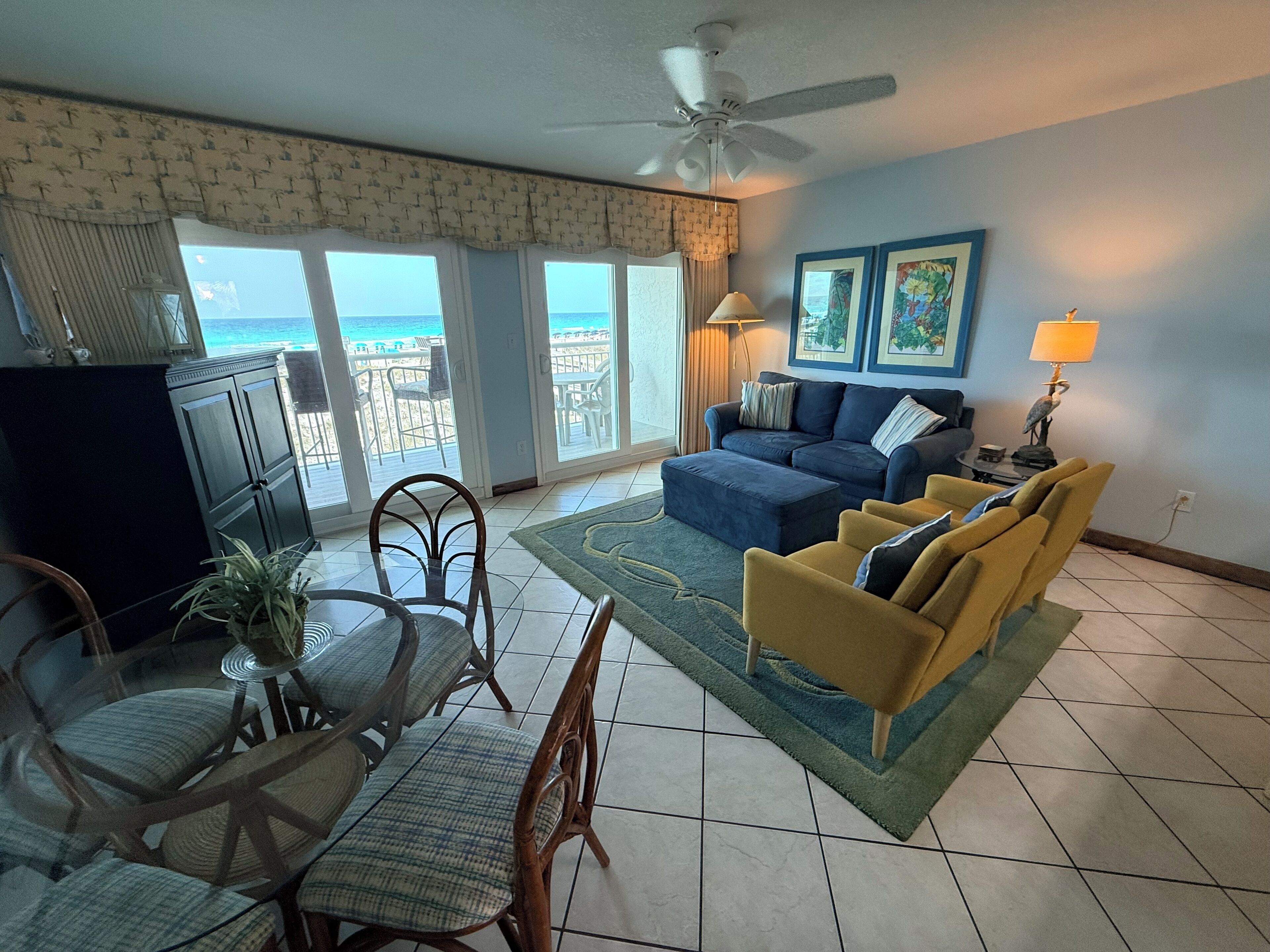 A living room with a view of the ocean, a ceiling fan, a dining area with chairs and a table, and a sofa.