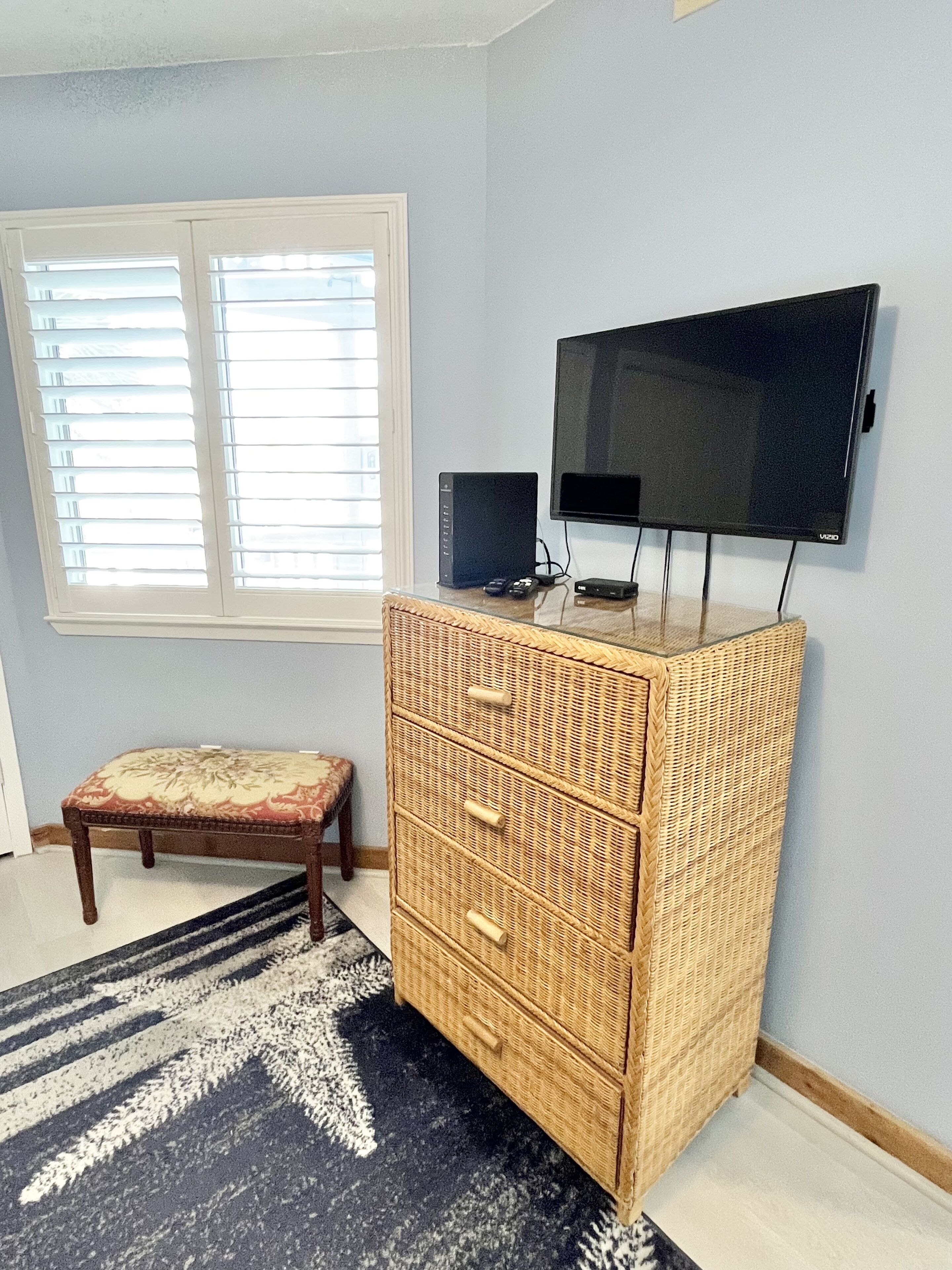 A room with a large window, a flat-screen TV mounted on the wall, a wicker dresser, and a patterned rug.