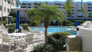 Zona de piscina con sillones de descanso y sombrillas, rodeada de palmeras y un edificio con balcones.