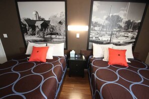 A hotel room with two beds, each with a patterned bedspread and a red pillow. There is a nightstand between the beds with a lamp and a small table. - Koreana Apartments No.2 (Decorah)