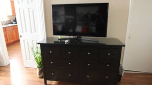 A dark wooden dresser with a flat-screen TV on top, a potted plant to the left, and a white door in the background. - Koreana Apartments No.2 (Decorah)