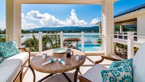 A balcony with a view of a beach and mountains, a round table with a coffee pot, and a seating area with patterned cushions. - 5 BDR LUX MODERN BEACHFRONT VILLA PRIVATE INFINITY POOL & OCEAN VIEWS STAFF (Montego Bay)