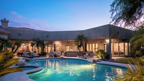 A pool area with lounge chairs and a bar, surrounded by palm trees and a building with large windows.