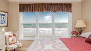 A bedroom with a large window overlooking the beach, a bed with patterned bedding, a chair, a lamp, and a framed picture on the wall. - A Villa Roja: Oceanfront Home with Incredible Views, Deck, Open Concept (Oak Island)