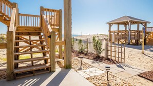 A wooden outdoor deck with a staircase leading to a covered area, a gazebo, and a sandy beach in the background. - A Villa Roja: Oceanfront Home with Incredible Views, Deck, Open Concept (Oak Island)