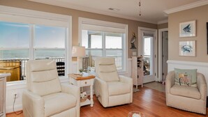 A spacious room with two white leather chairs, a wooden table, and a ceiling fan. Large windows offer a view of the ocean. - A Villa Roja: Oceanfront Home with Incredible Views, Deck, Open Concept (Oak Island)