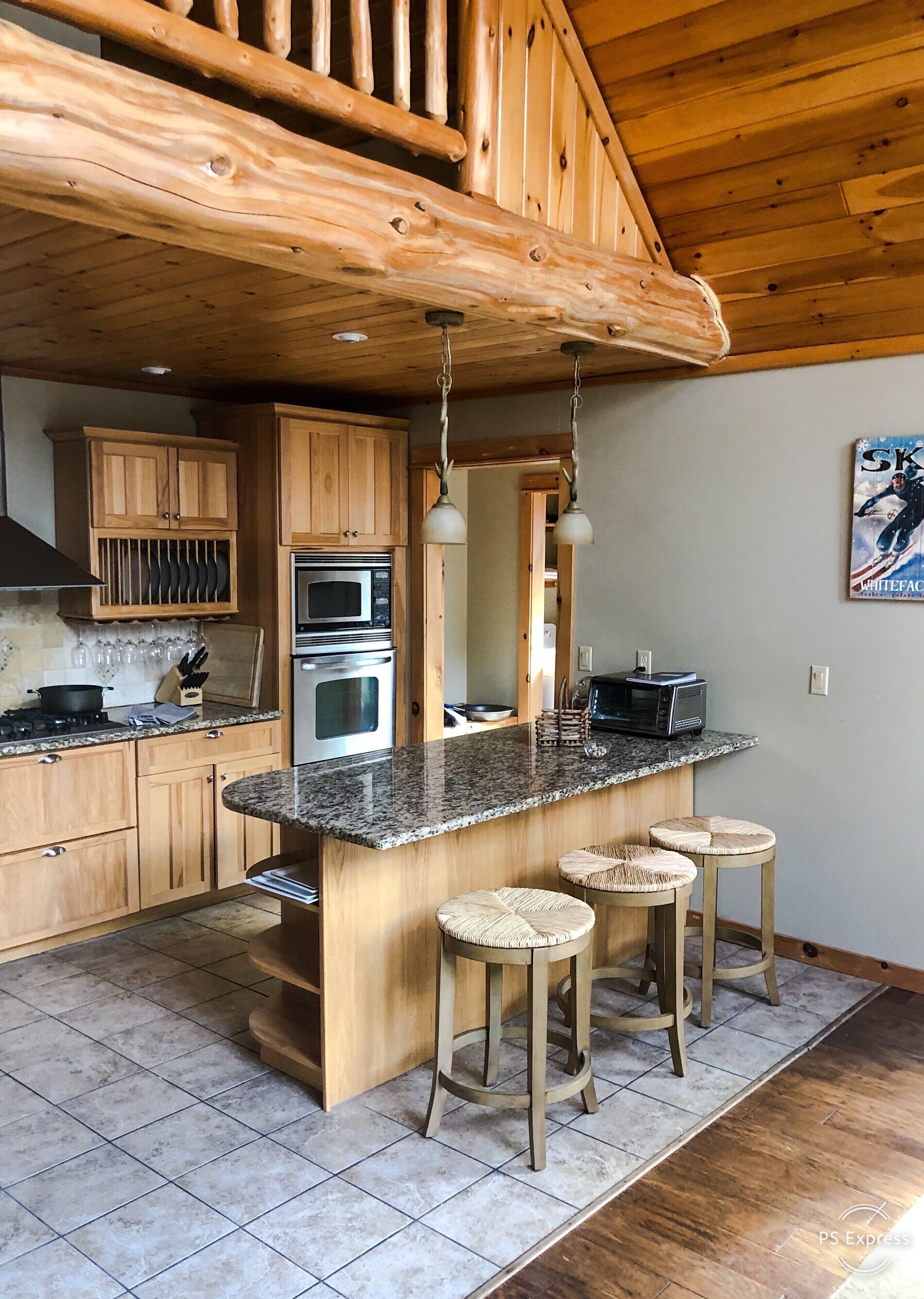 Une cuisine dotée de meubles en bois, de plans de travail en granit, d’un four encastré et d’un coin bar avec des tabourets.