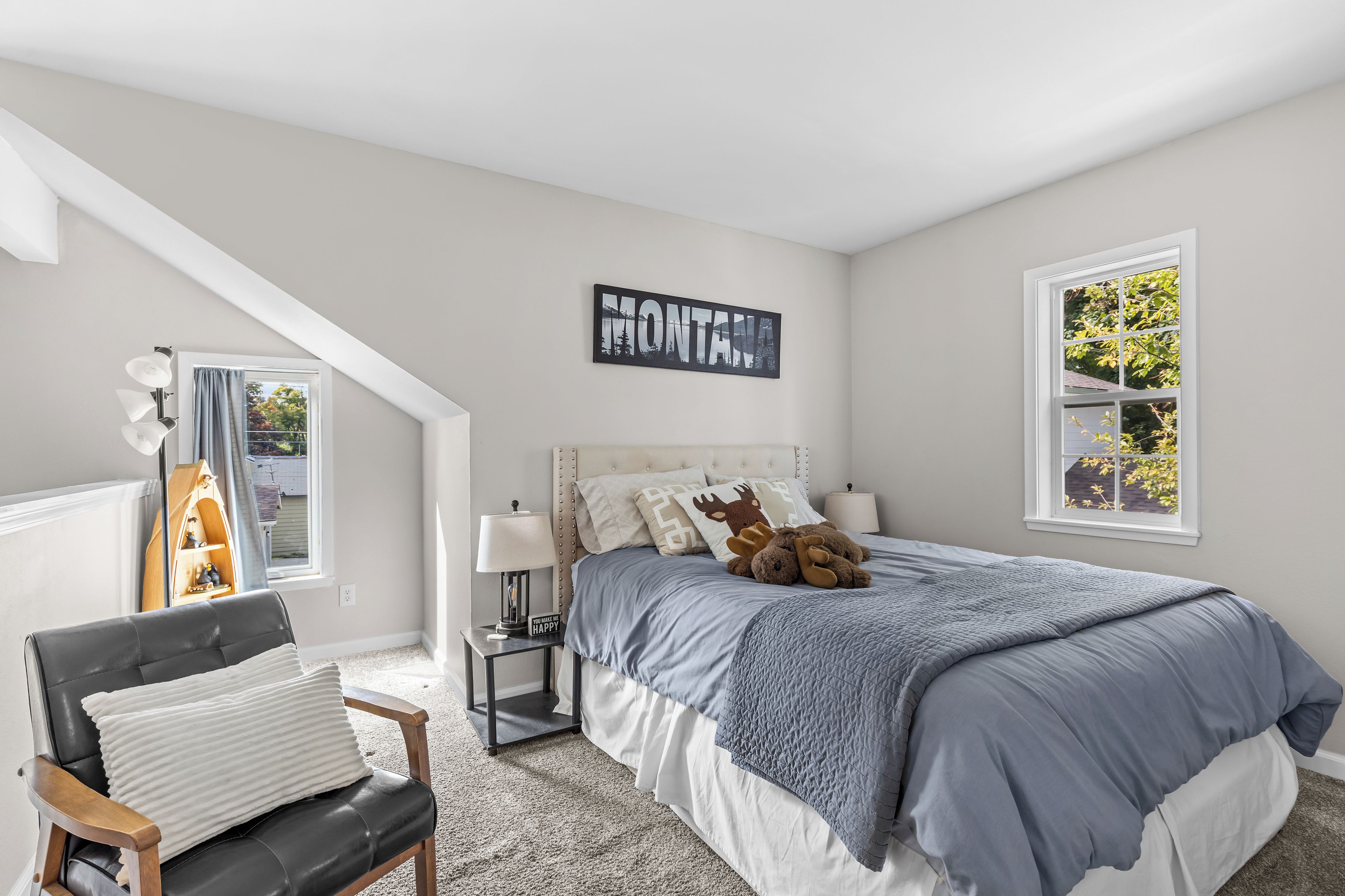 A bedroom with a bed, a chair, a nightstand, a lamp, a window, and a wall-mounted sign.