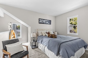 Une chambre à coucher comprenant un lit, une chaise, une table de chevet, une lampe, une fenêtre et un panneau fixé au mur.