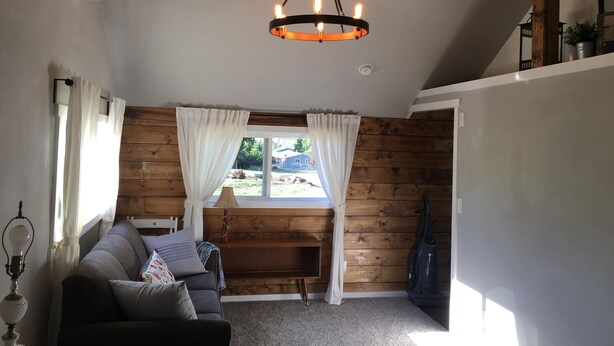 A cozy living room with a wooden wall, a sofa, a window with curtains, and a hanging light fixture. - Quiet Cottage in the Country (Cove)