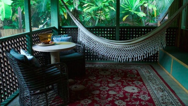 Un fauteuil suspendu, une table ronde et un tapis à motifs dans une pièce offrant une vue sur une végétation luxuriante.
