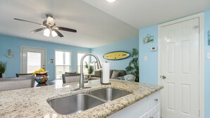 A kitchen with granite countertops, stainless steel sink, ceiling fan, and a surfboard decoration on the wall. - Beachside Condo | Heated Pool, Hot Tub & Dune Views (Corpus Christi)