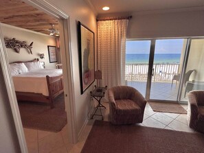 Une chambre avec un lit, une chaise, une petite table et un tableau. Il y a un balcon avec vue sur la plage.