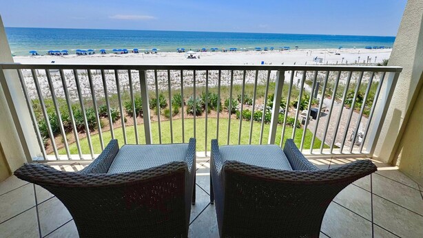 Un balcon avec des fauteuils en osier donnant sur une plage parsemée de parasols et où se trouvent des gens.