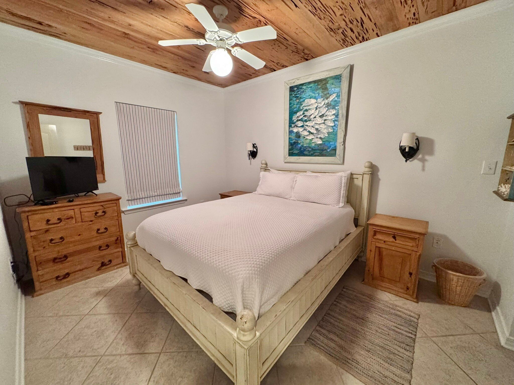 A bedroom with a wooden ceiling, a bed with a white bedspread, a wooden dresser, a television on the dresser, a window with blinds, and a painting on the wall.
