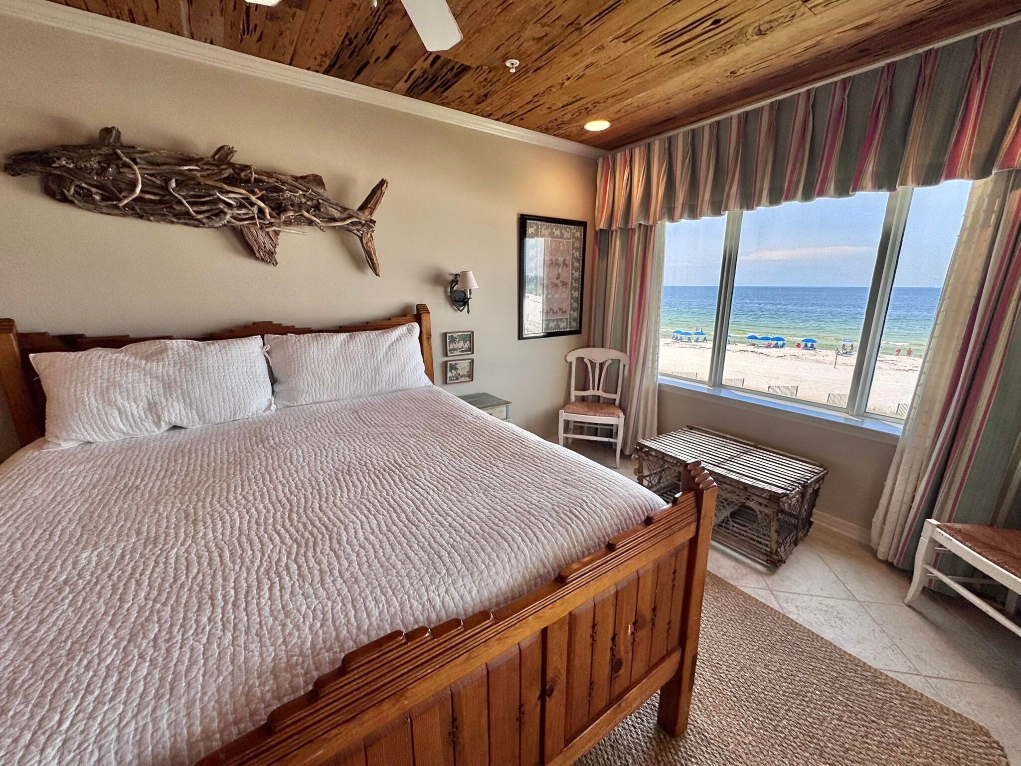 Ein Schlafzimmer mit einem großen Bett, einem Holzkopfende, einer Holzzielung, Blick auf den Strand, einem Sessel und einem kleinen Tisch.