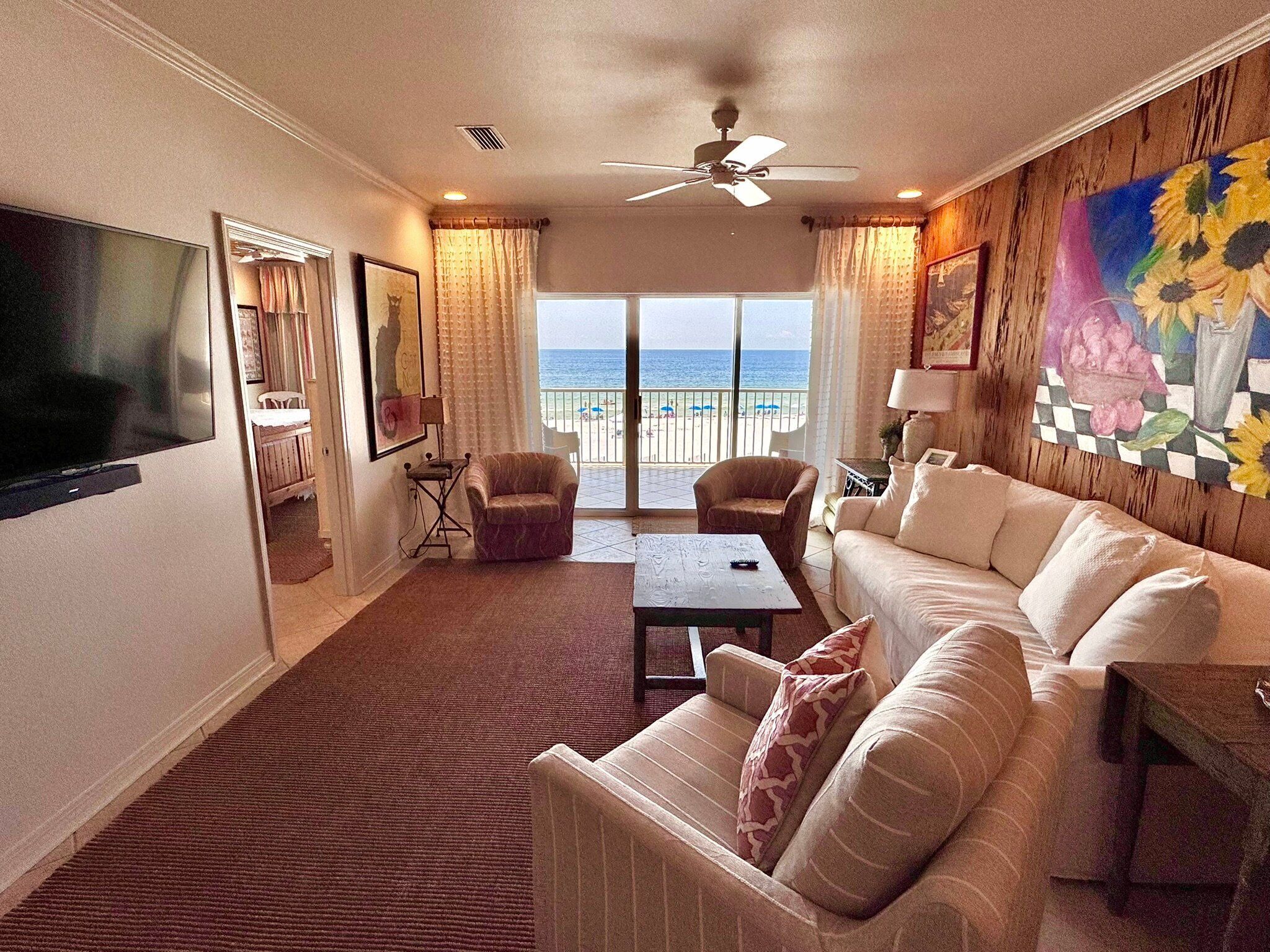 A spacious living room with a large flat-screen TV, a ceiling fan, and a view of the beach.