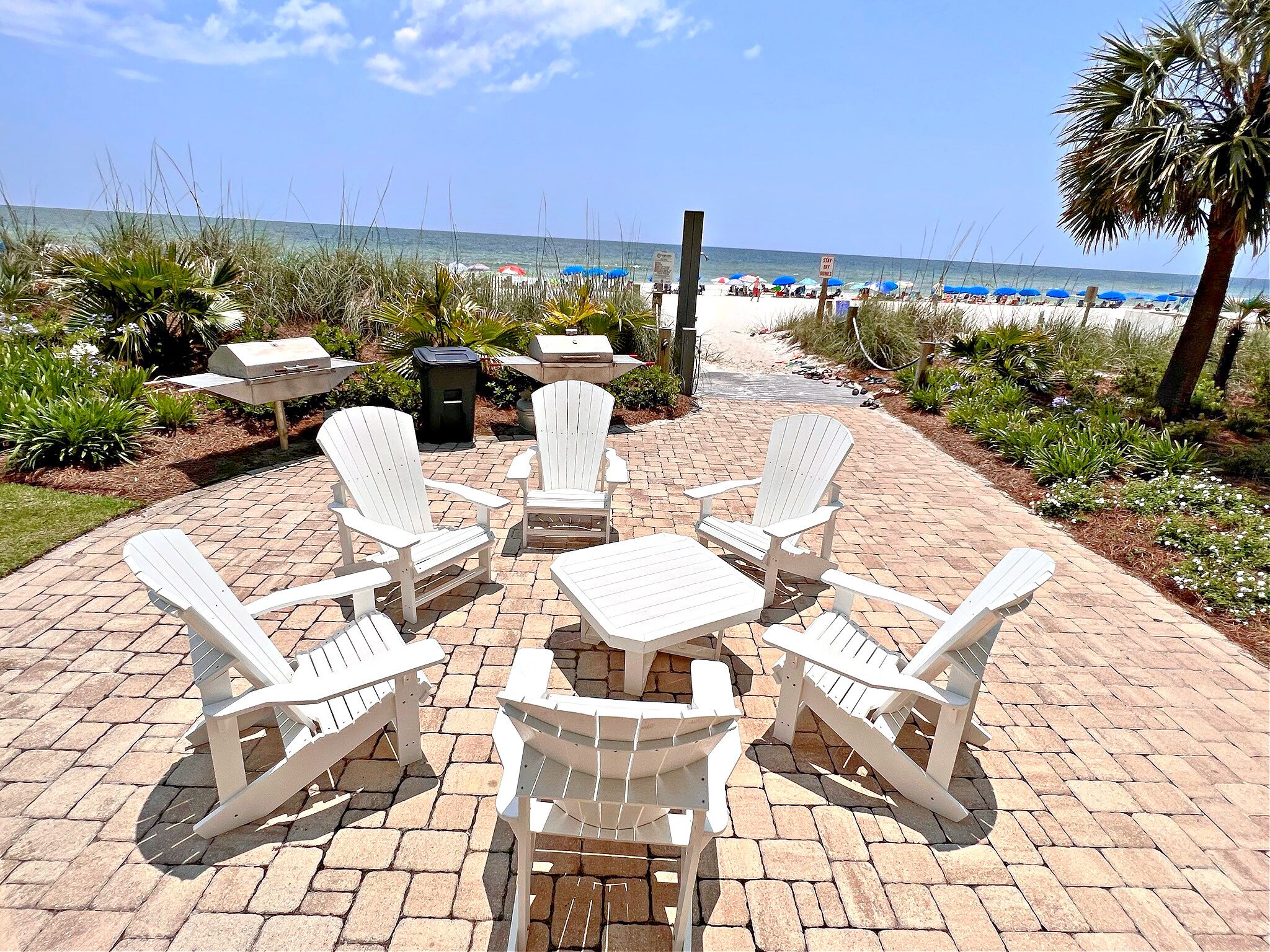 A patio with Adirondack chairs and a table, overlooking a beach with umbrellas and palm trees.