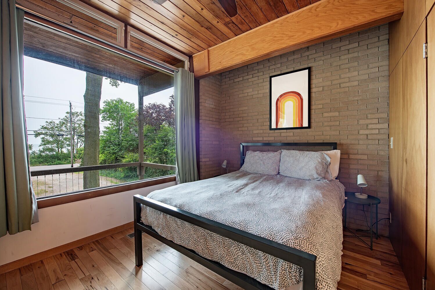 A bedroom with a large window, a bed with patterned bedding, a brick wall, and a framed artwork.