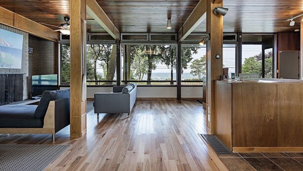 A modern interior with wooden floors, a sofa, a TV, and large windows overlooking a body of water. - GLASS HOUSE 5 BR Private Lake Erie Beach Great for Summer, Fall, Winter (Sandusky)
