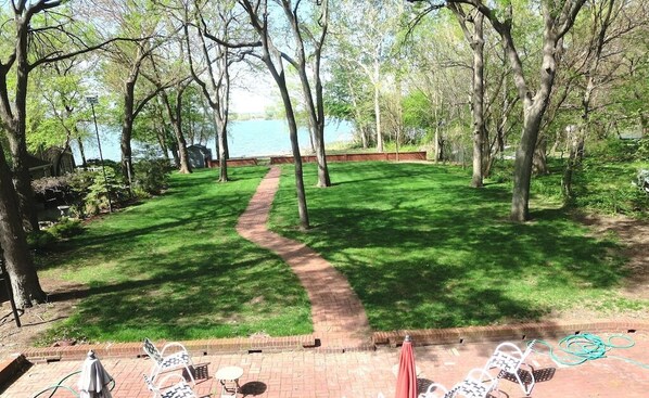 A grassy backyard with a brick pathway leading to a body of water, surrounded by trees. - GLASS HOUSE 5 BR Private Lake Erie Beach Great for Summer, Fall, Winter (Sandusky)