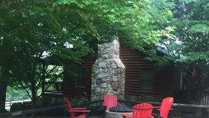 A log cabin with a stone fireplace, surrounded by a wooden deck and red Adirondack chairs.