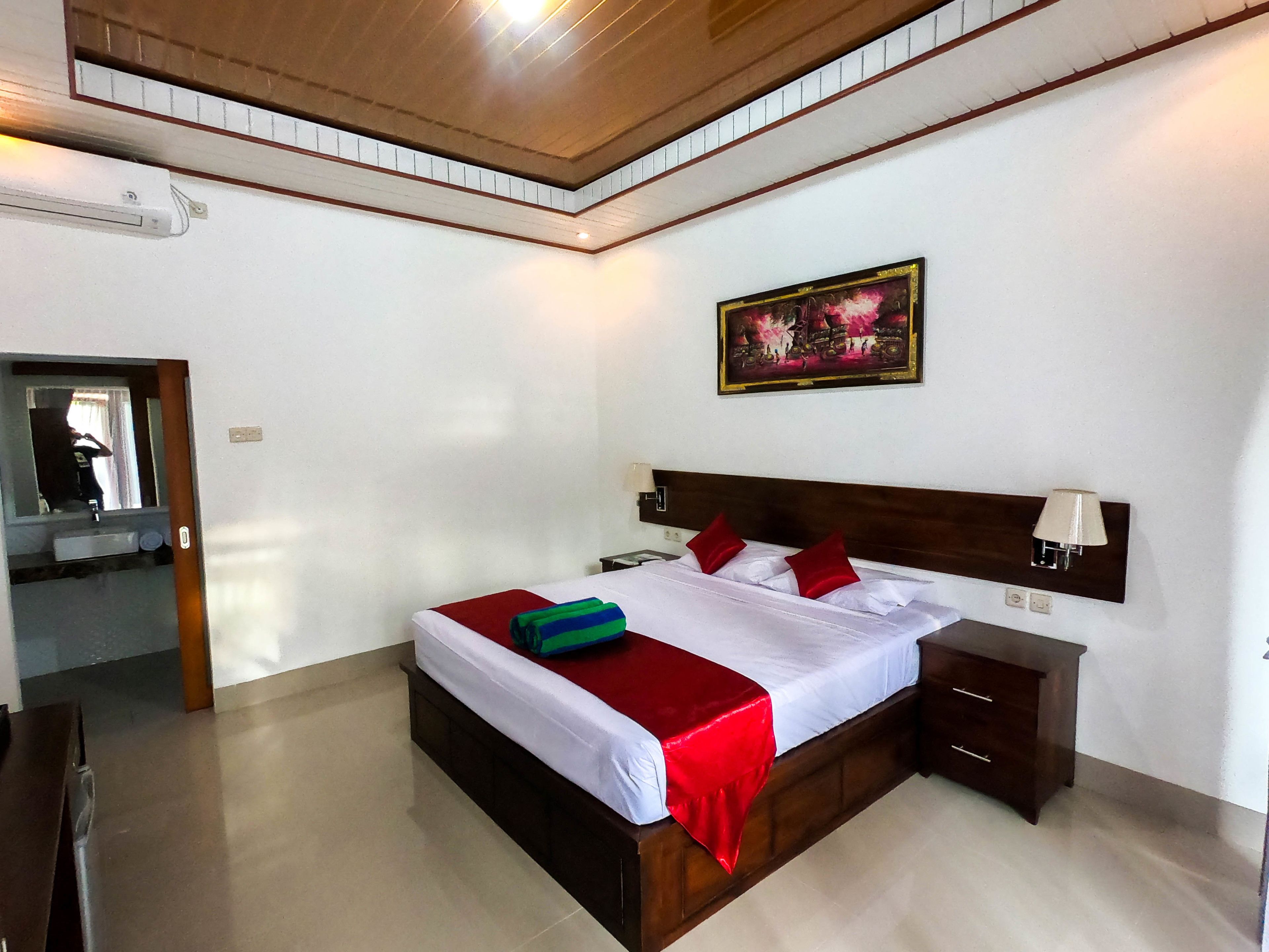 Habitación de hotel con techo de madera, una cama grande con ropa de cama roja y blanca, una mesita de noche, una lámpara de pared y un cuadro sobre la cama.