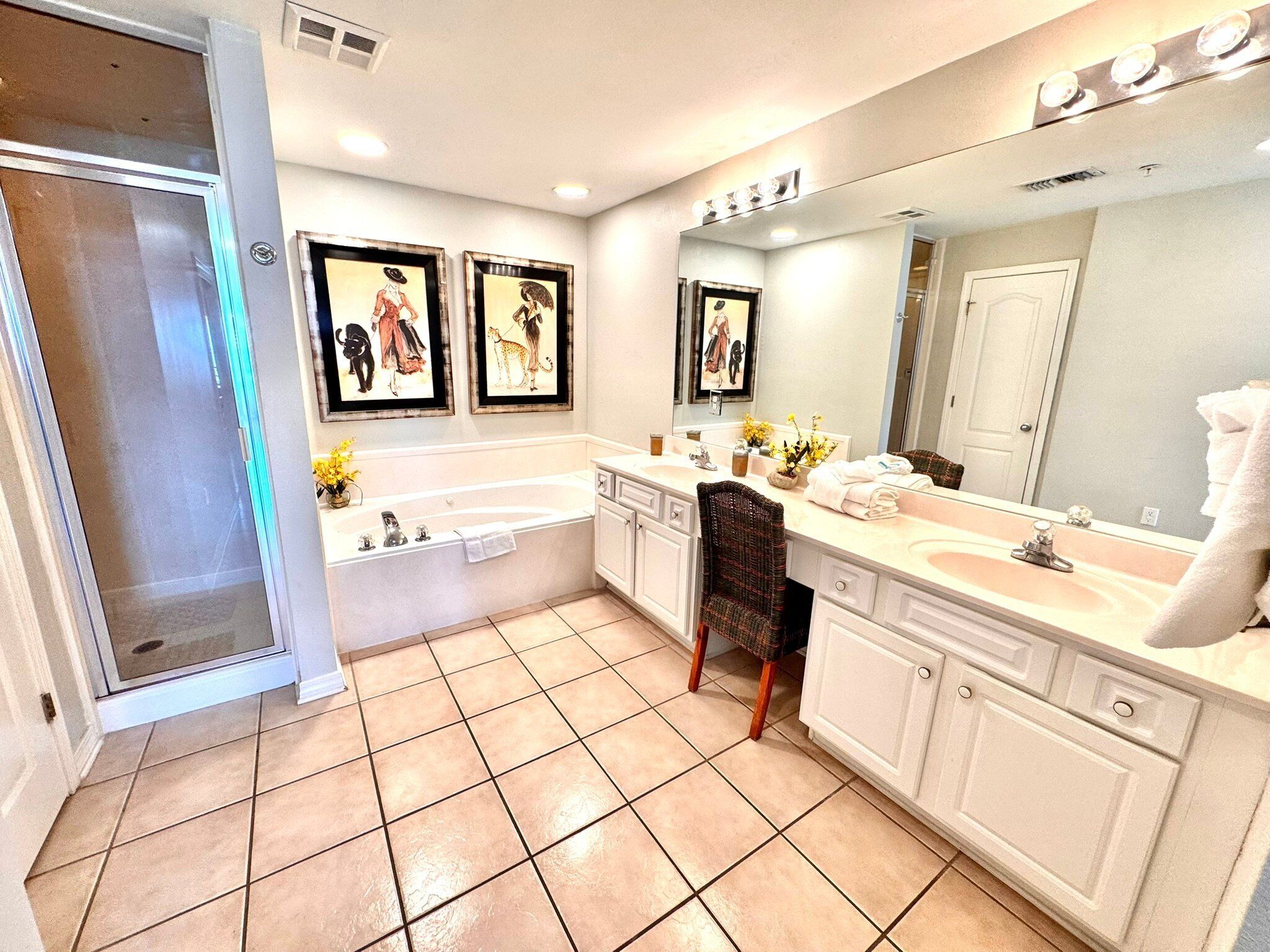A bathroom with a large bathtub, a double sink vanity, and a chair. There are framed pictures on the wall and a large mirror above the sink.