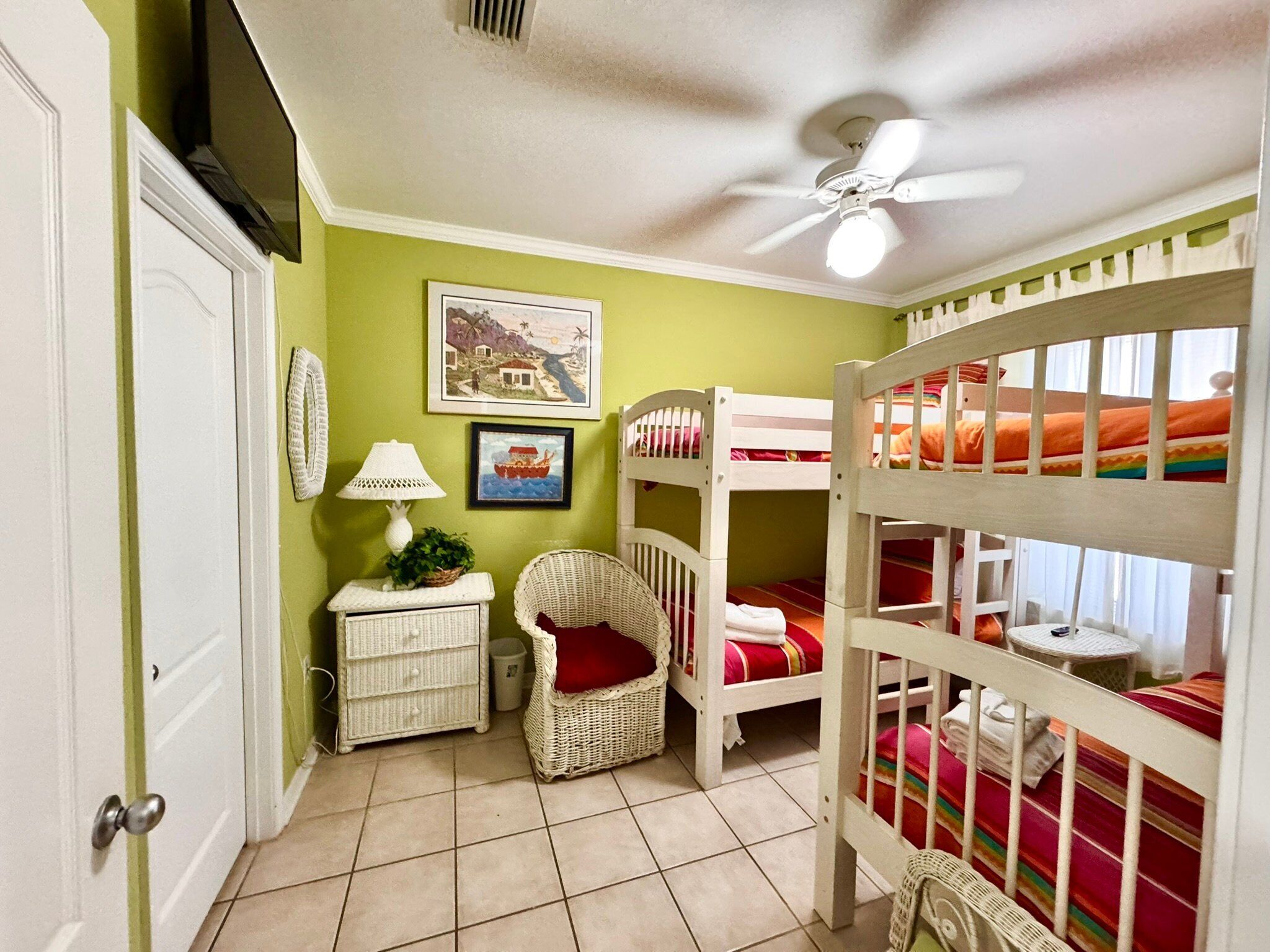 Une chambre d’enfant avec des lits superposés, un ventilateur de plafond, une chaise en osier et une petite table.