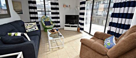 Un salon avec un canapé, un fauteuil, une télévision et un balcon agrémenté de rideaux à rayures.