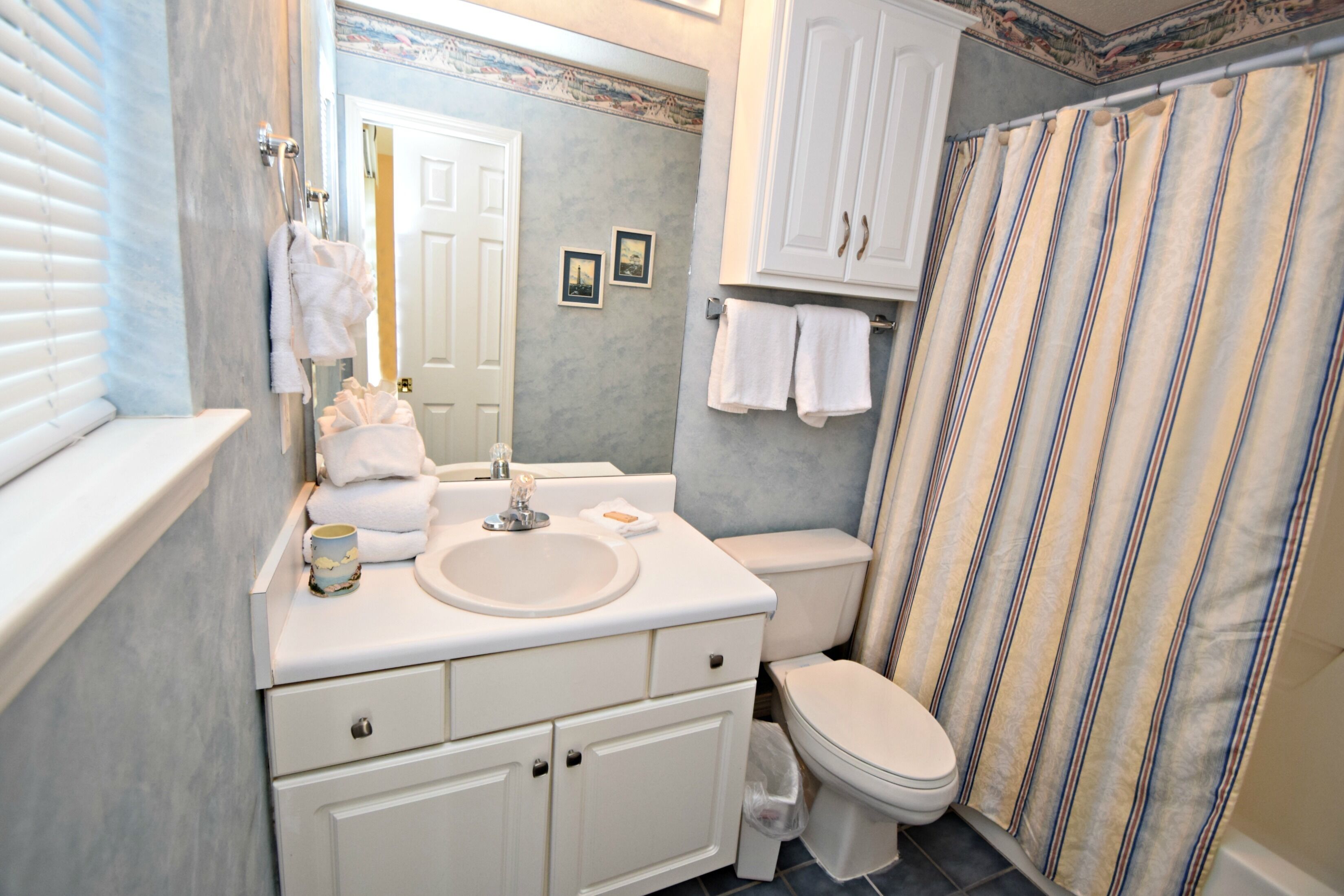 Une salle de bain avec un lavabo blanc, des toilettes et un rideau de douche. Des serviettes sont accrochées au mur, et il y a une petite poubelle.