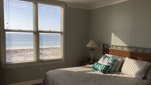 Ein Schlafzimmer mit großem Fenster, einem Bett mit Kopfteil, einem Nachttisch und einer Lampe.