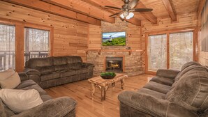 Ein Blockhaus-Interieur mit Kamin aus Stein, Ledersofas und einem darüber angebrachten Fernseher.
