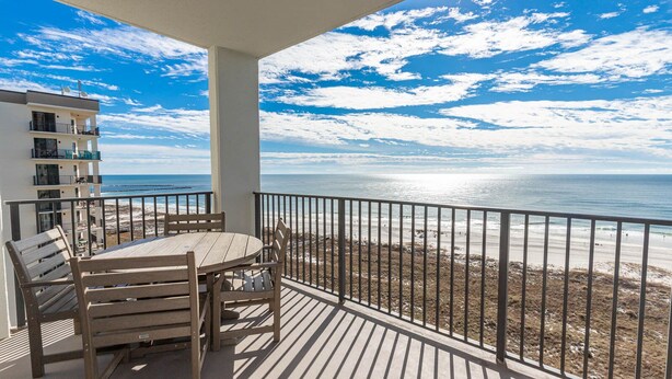 Un balcon avec une table et des chaises en bois, donnant sur une plage sous un ciel dégagé.