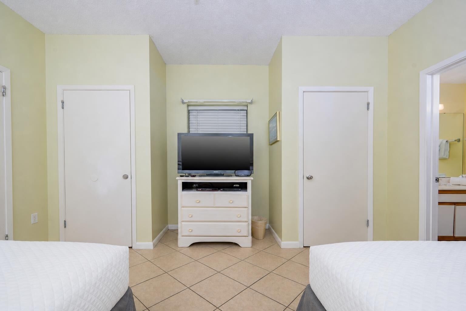 A hotel room with two beds, a TV on a stand, a small window with blinds, and a door on the right.