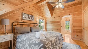 A cozy bedroom with a wooden ceiling, a bed with a floral bedspread, a nightstand with a lamp, and a ceiling fan.