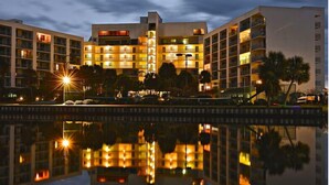 Ein mehrstöckiges Hotel mit Balkonen, einem spiegelnden Pool und wolkenlosem Himmel.