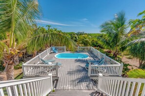 Una terraza de madera con piscina, rodeada de palmeras y una valla blanca.
